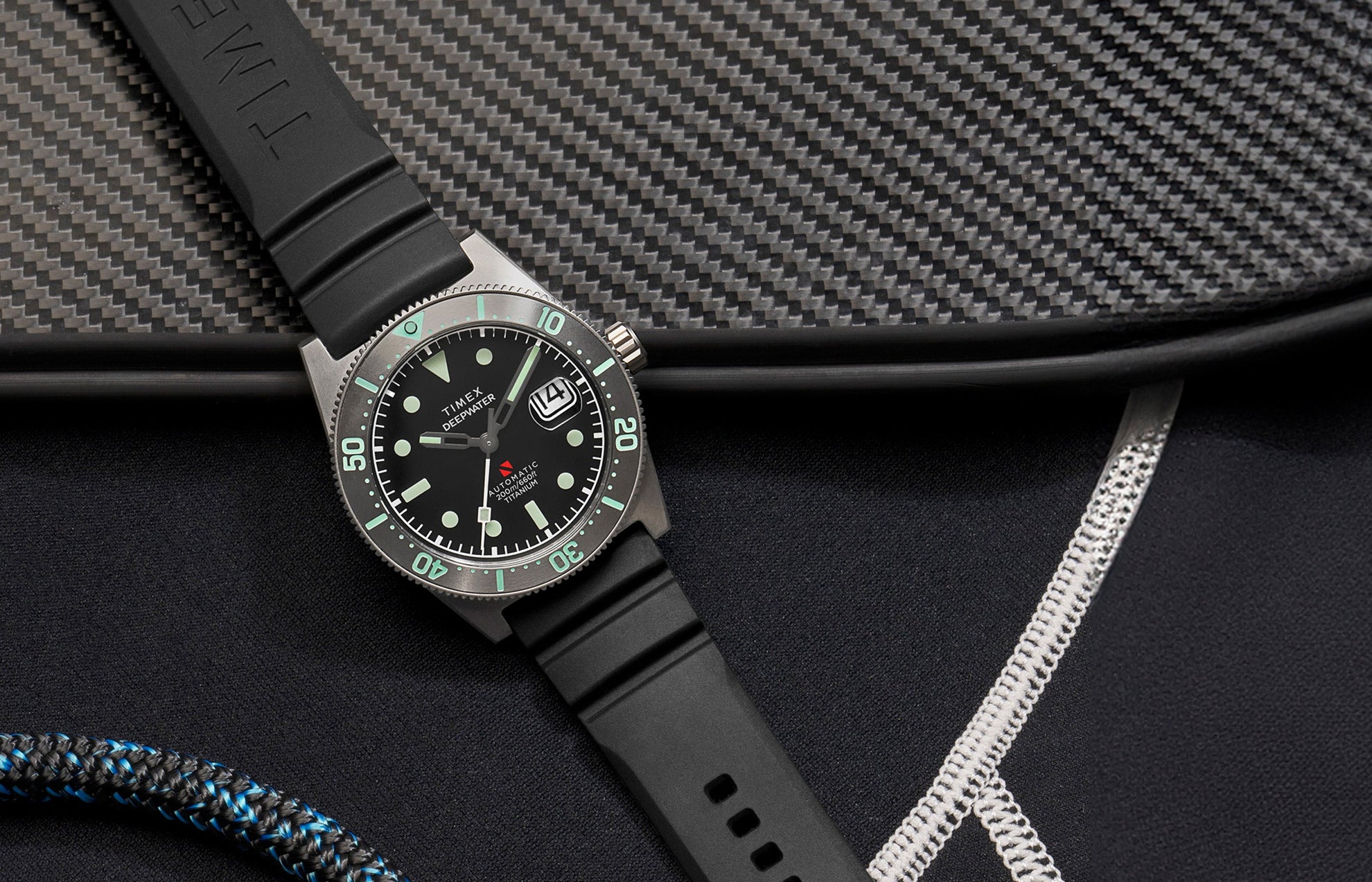 Timex Deepwater Reef 200 Titanium Automatic watch with black dial and luminous markers, displayed on a black rubber strap beside carbon fiber gear and diving equipment.