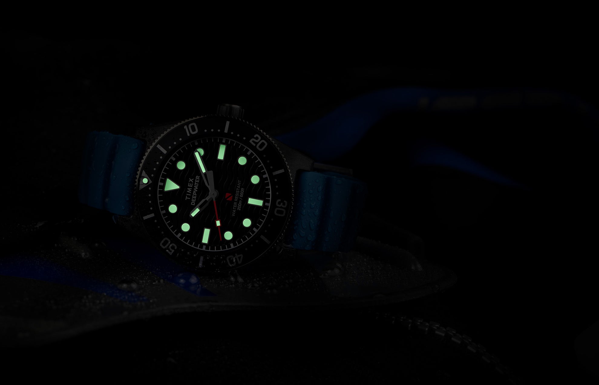 A Timex Deepwater dive watch with a black dial and luminous green markers glowing in the dark, featuring a blue silicone strap and water droplets on the case, emphasizing its water-resistant design.
