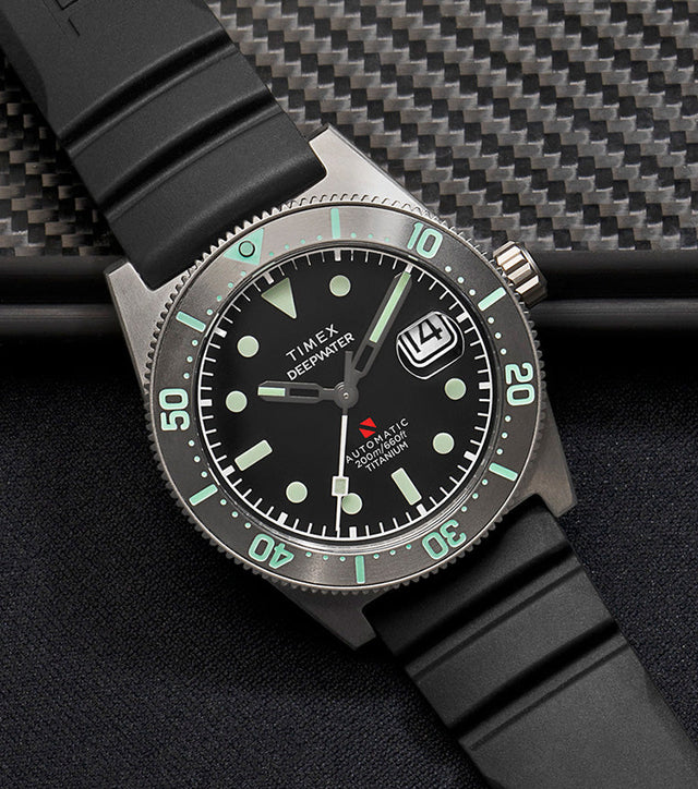 Timex Deepwater Reef 200 Titanium Automatic watch with black dial and luminous markers, displayed on a black rubber strap beside carbon fiber gear and diving equipment.
