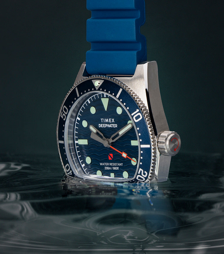 Timex Deepwater Reef 200 watch with blue dial and matching blue rubber strap, half-submerged in rippling water against a dark background, highlighting its aquatic performance design.