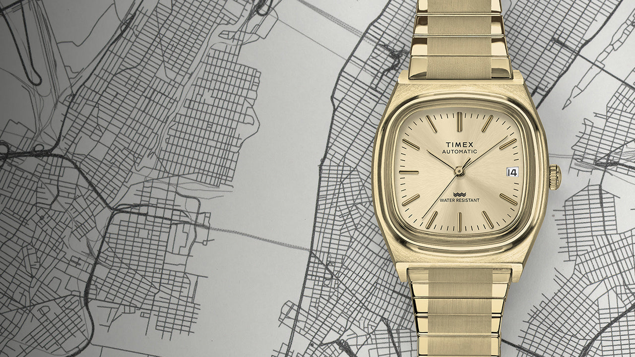 Full shot of the Timex® Automatic 1983 E Line Reissue in gold-tone stainless steel positioned over a city map. The image highlights the retro-inspired cushion case, sunburst champagne dial, and integrated bracelet design.