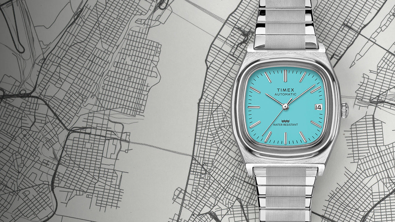 Full view of the Timex® Automatic 1983 E Line Reissue in stainless steel with turquoise dial laid over a grayscale city map. The image emphasizes the retro-inspired square case design and integrated bracelet.
