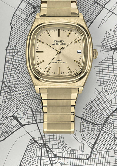 Full shot of the Timex® Automatic 1983 E Line Reissue in gold-tone stainless steel positioned over a city map. The image highlights the retro-inspired cushion case, sunburst champagne dial, and integrated bracelet design.