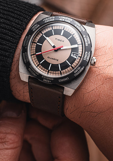 Lifestyle image of the Q Timex® 1972 World Time watch on wrist, displaying the bold tan-and-black world time dial, brushed steel case, red seconds hand, and brown leather strap paired with a dark sweater.