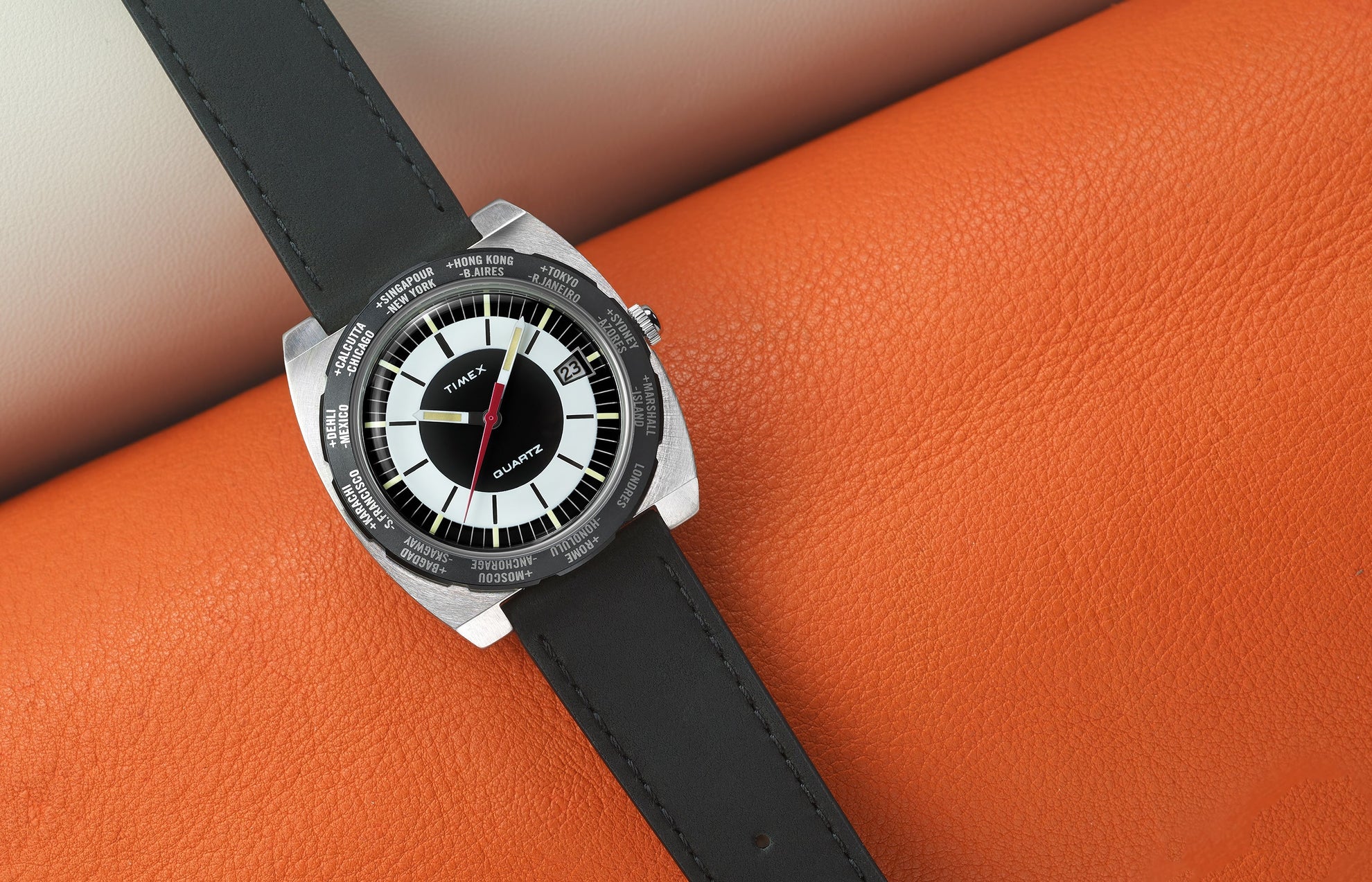 Top-down view of the Timex World Time Reissue watch with a black leather strap, silver brushed case, and black-and-white dial featuring a red seconds hand. The bezel shows world cities like Singapore, Hong Kong, and Tokyo. The watch rests on an orange textured leather surface.