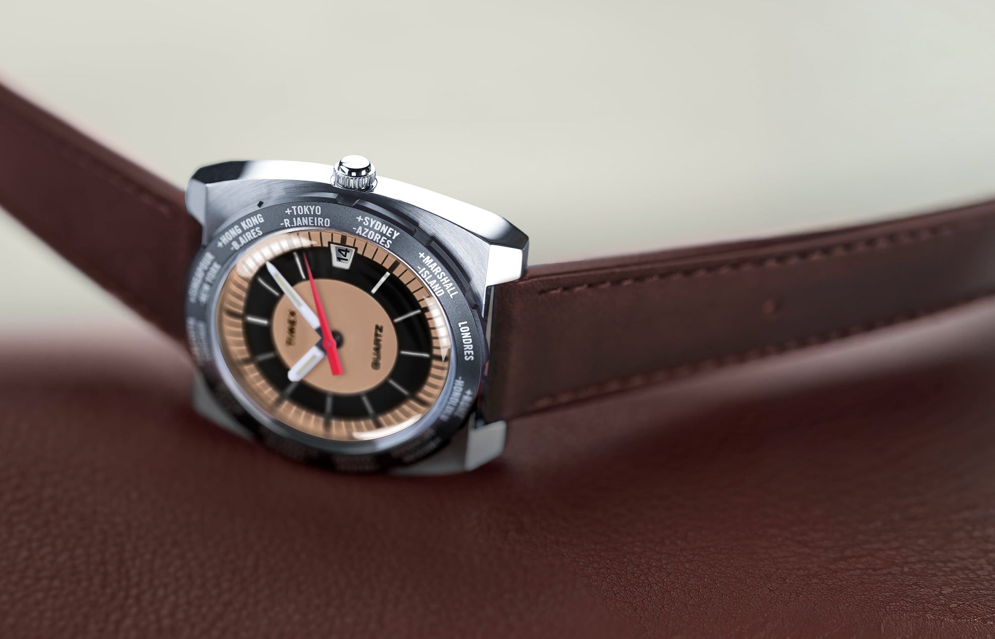 Angled view of the Q Timex® 1972 World Time watch resting on brown leather, showing the brushed stainless-steel case, brown leather strap, and tan-and-black world time dial with a red seconds hand.