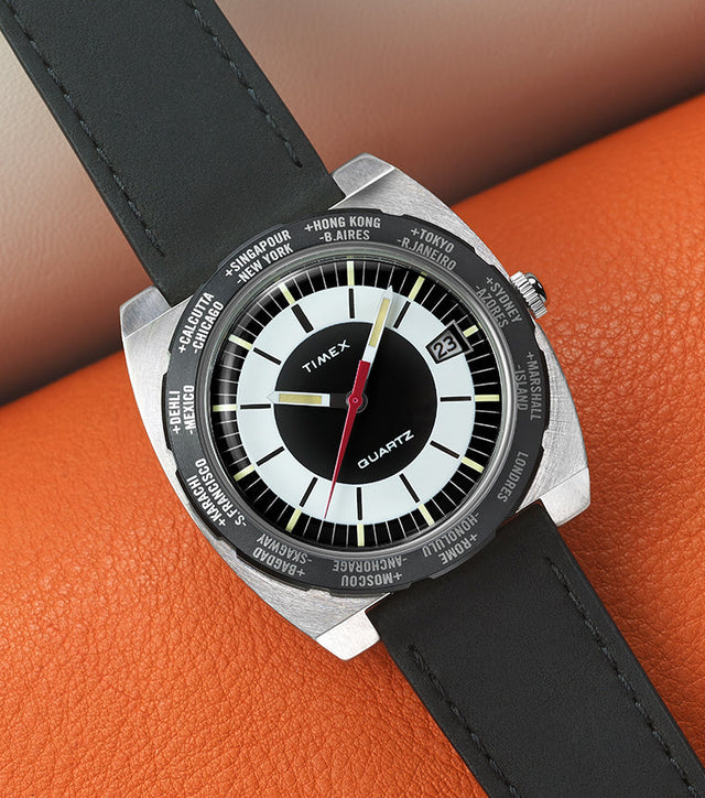 Top-down view of the Timex World Time Reissue watch with a black leather strap, silver brushed case, and black-and-white dial featuring a red seconds hand. The bezel shows world cities like Singapore, Hong Kong, and Tokyo. The watch rests on an orange textured leather surface.
