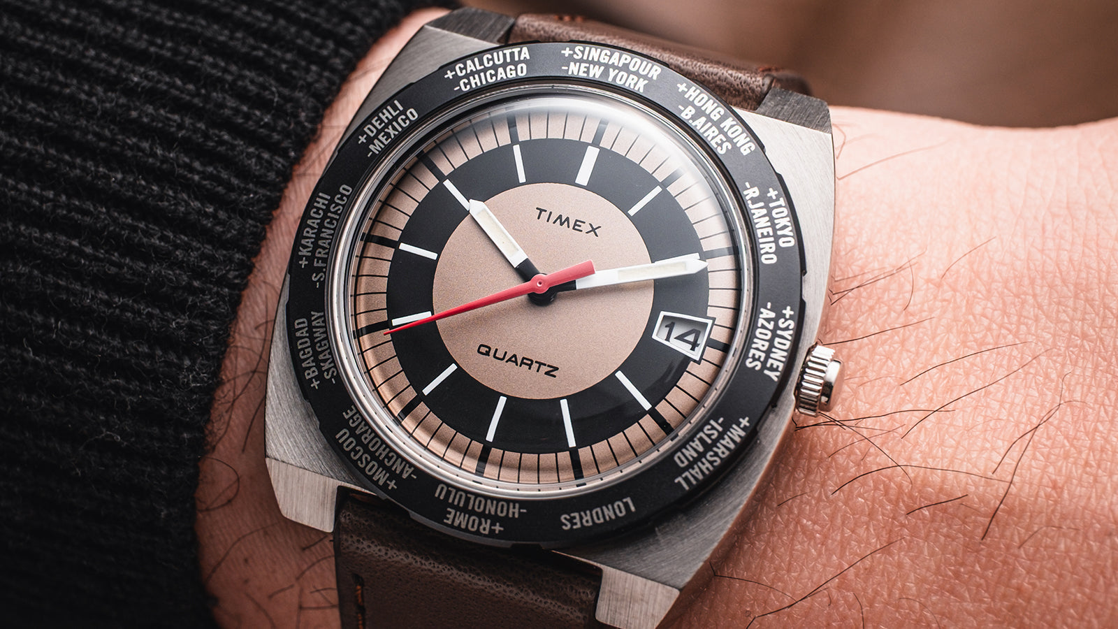 Wrist shot of the Q Timex® 1972 World Time watch showing the retro square case, tan-and-black dial with red seconds hand, and rotating bezel engraved with global city names.