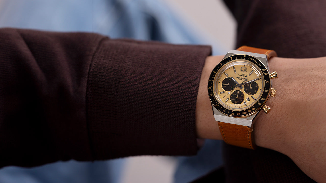 Q Timex Chronograph with a gold dial, black sub-dials, and tan leather strap worn on wrist, paired with a brown sweater and blue shirt.