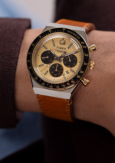 Q Timex Chronograph with a gold dial, black sub-dials, and tan leather strap worn on wrist, paired with a brown sweater and blue shirt.