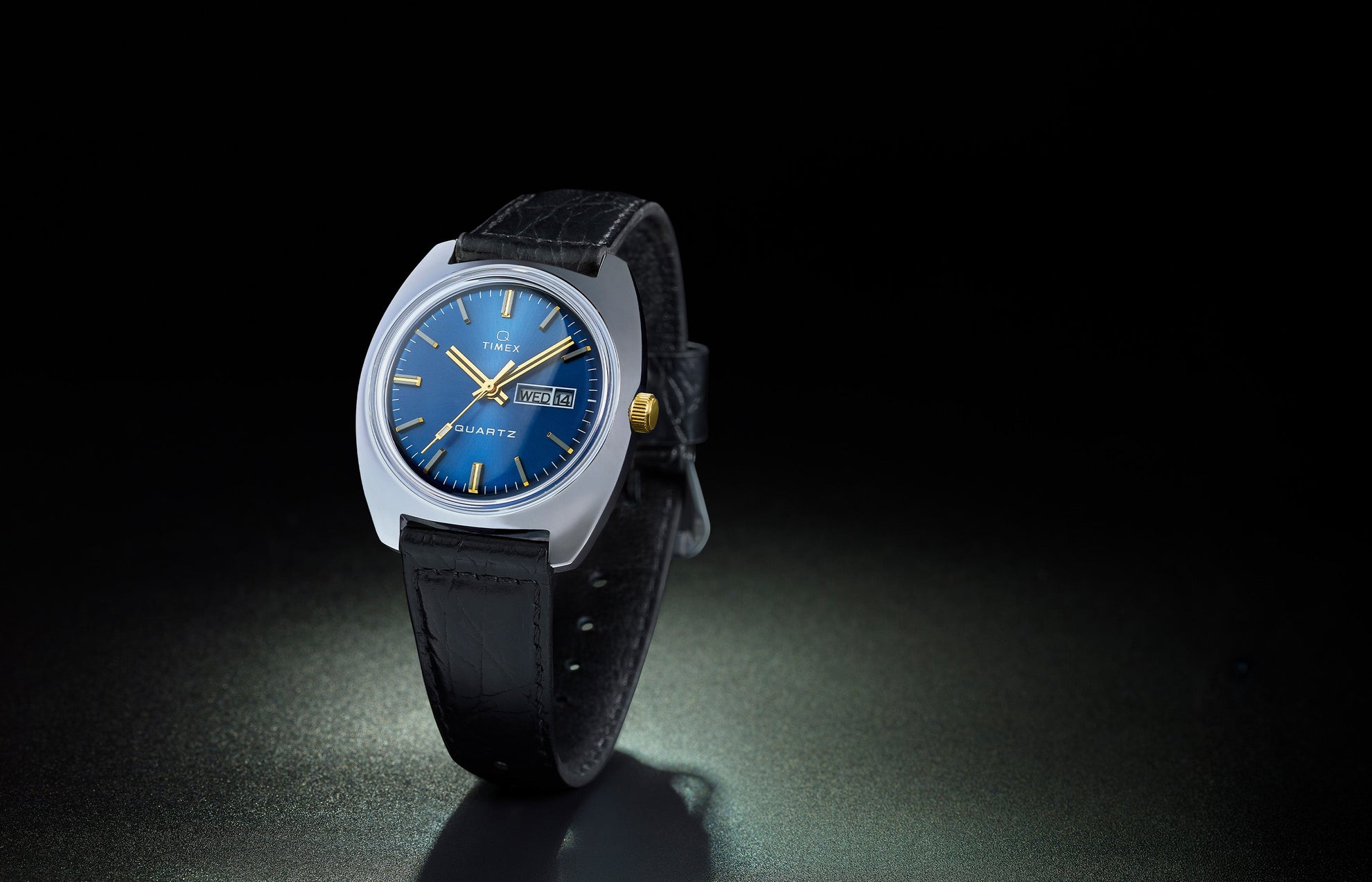 Q Timex® 1978 Day/Date watch with a blue sunray dial, gold-tone hands, and a black leather strap, displayed against a dark background with a subtle spotlight effect.