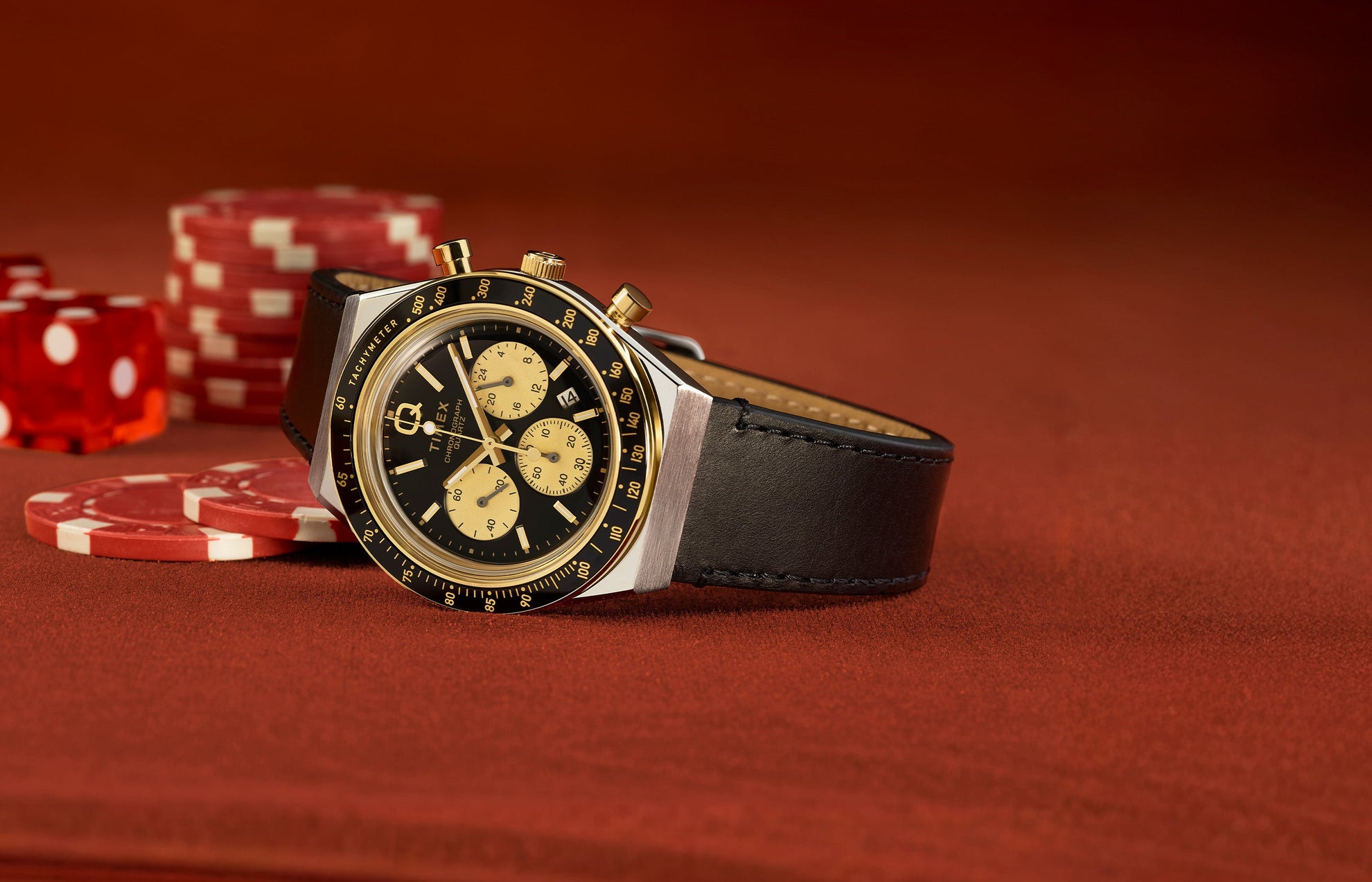 Q Timex Chronograph with a black dial and gold-tone sub-dials on a black leather strap, resting on a poker table with red chips and dice in the background.