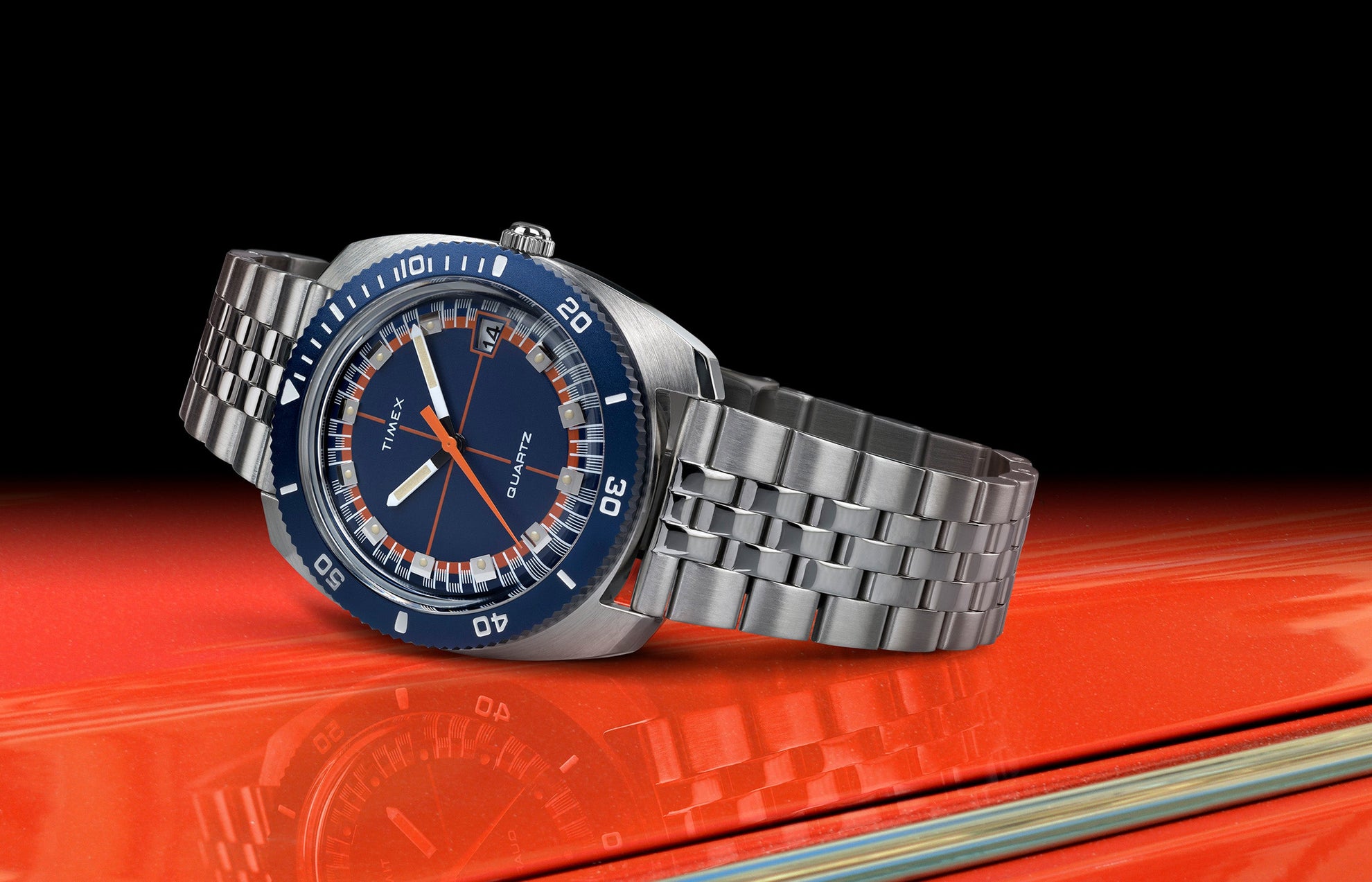 Q Timex® 1971 Velocity watch with a blue dial, orange accents, and stainless steel bracelet, displayed on a glossy red surface with a black background.