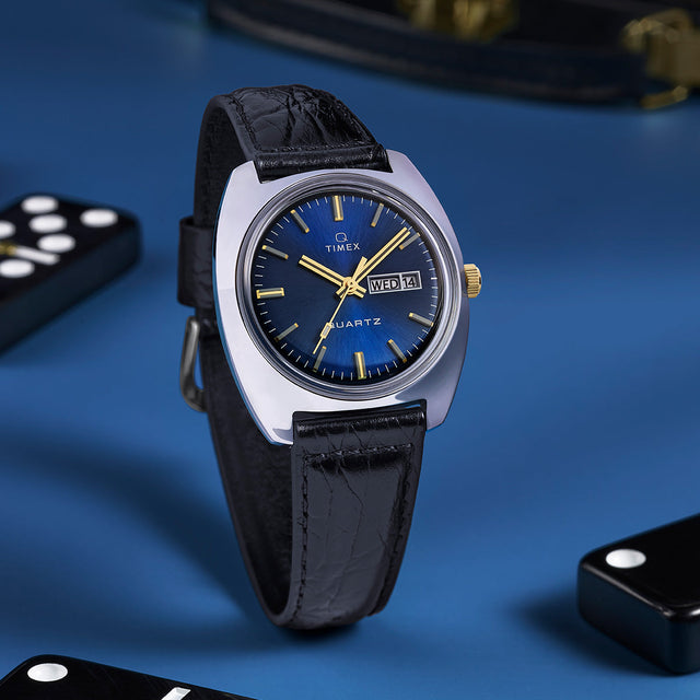 Q Timex® 1978 Day/Date watch standing upright on a blue surface with black dominoes, showcasing its polished case, blue dial, and gold-tone crown and markers.