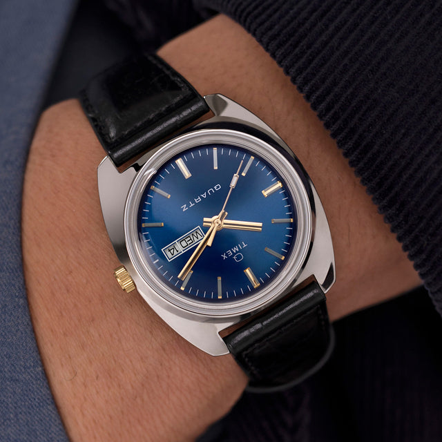 Q Timex® 1978 Day/Date watch worn on the wrist, showing the blue dial, gold-tone details, and black leather strap against a dark sleeve.