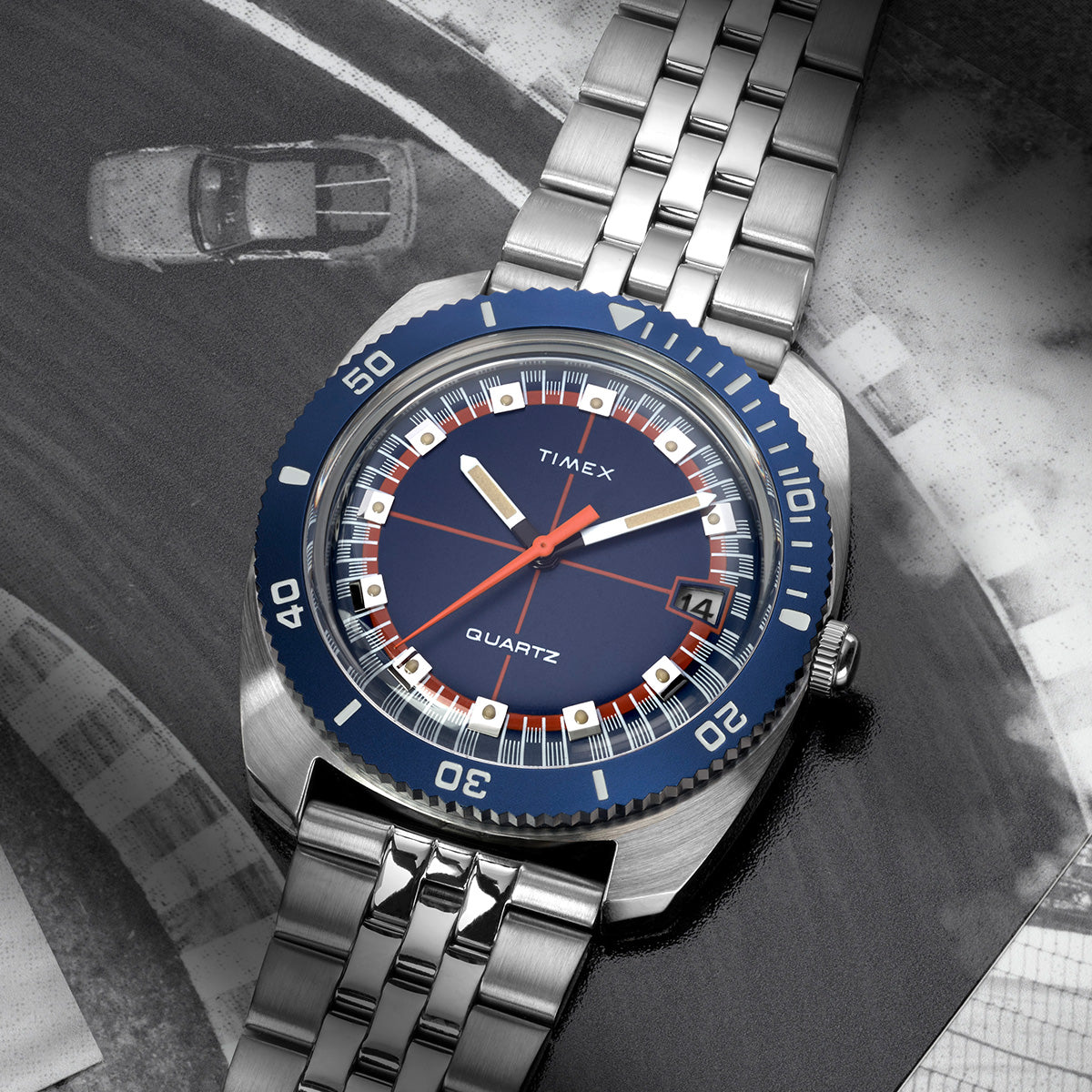Q Timex® 1971 Velocity watch with a blue dial and silver bracelet, placed over a black-and-white racing track photo, emphasizing its vintage motorsport inspiration.