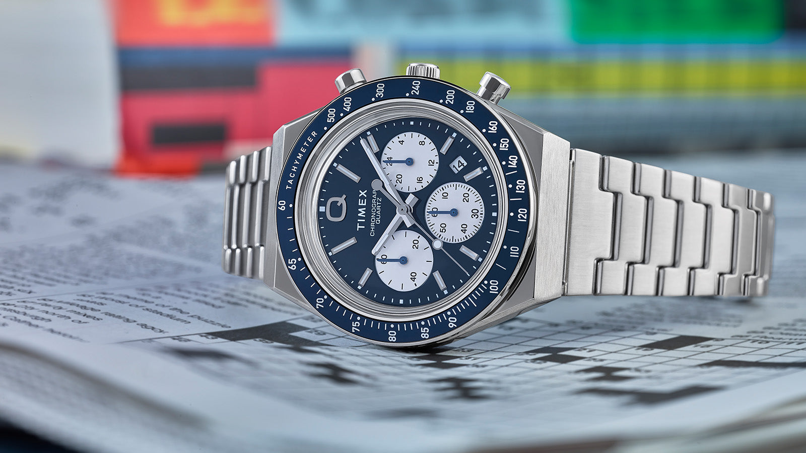 Q Timex Chronograph with a blue dial, white sub-dials, and stainless steel bracelet resting on a folded newspaper with colorful pages in the background.