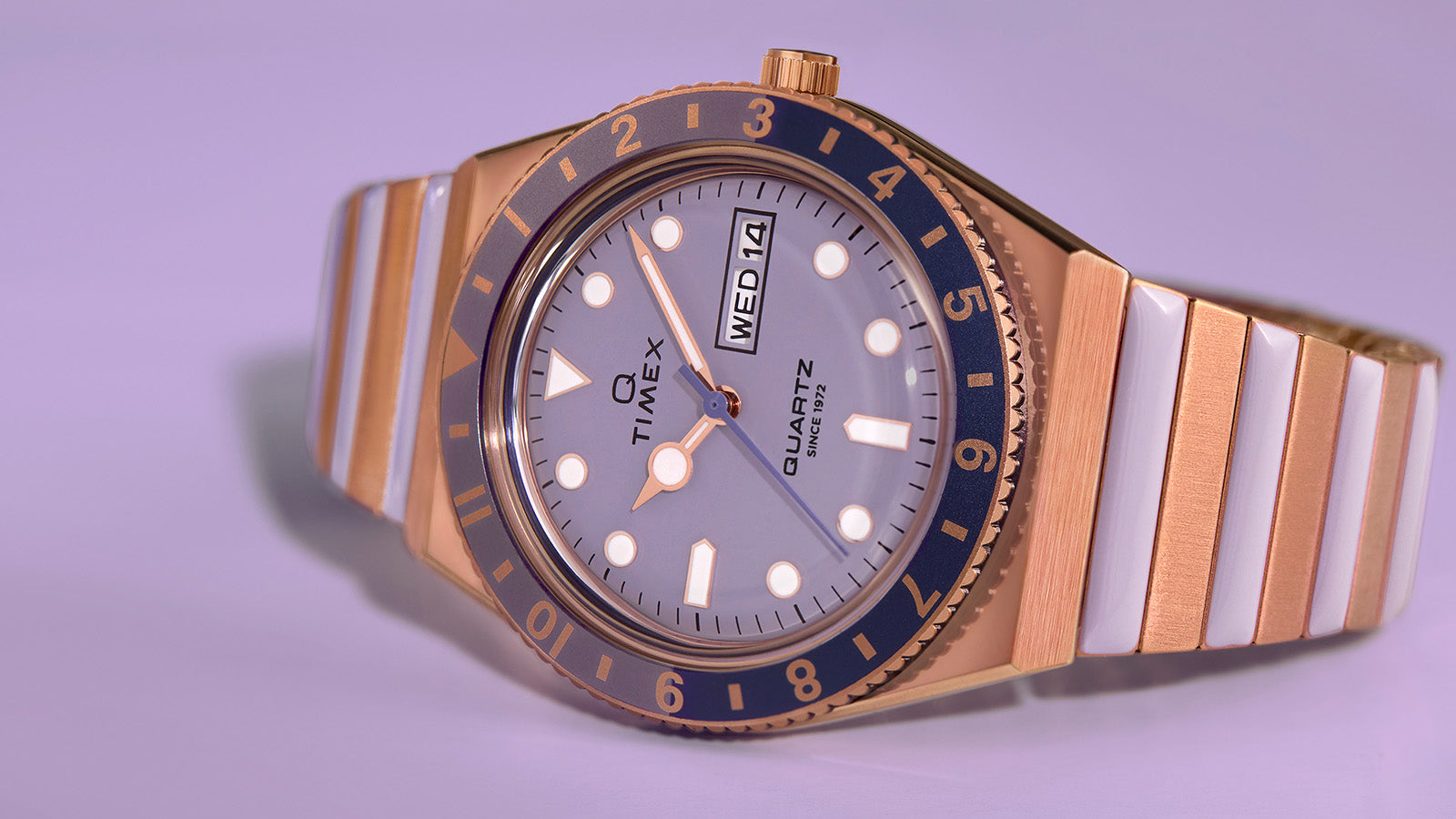 Close-up of the Q Timex® showing its lilac dial, blue and gold-tone bezel, and day-date window at 3 o’clock, with a rose gold-tone bracelet and polished finish.