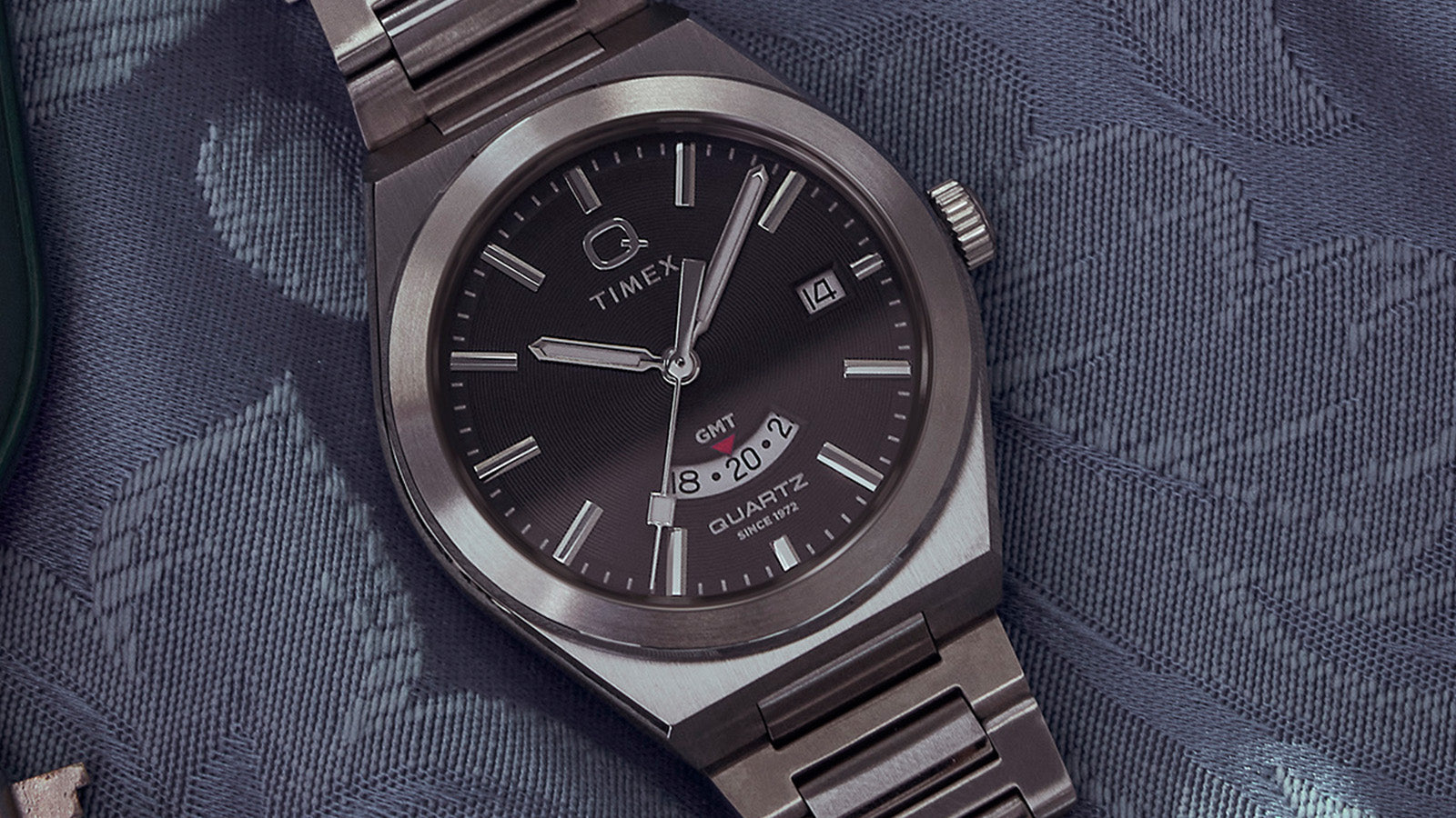 Detailed flat lay of the Q Timex® Continental GMT on blue fabric, focusing on the dial’s fine texture, sleek hands, and brushed metal bracelet links.