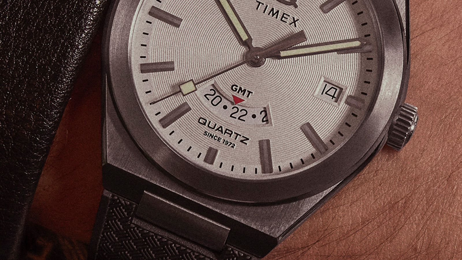 Macro shot of the Q Timex® Continental GMT dial showing fine circular texture, GMT subdial with red arrow indicator, and polished stainless-steel hands.