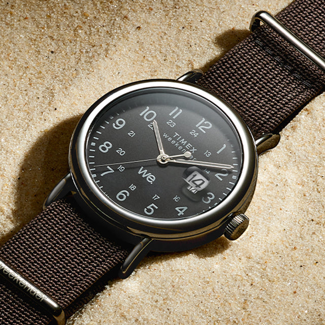 Timex Weekender watch with black dial and black fabric strap resting on sand, highlighting its silver-tone case and date window.