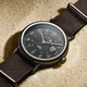 Timex Weekender watch with black dial and black fabric strap resting on sand, highlighting its silver-tone case and date window.