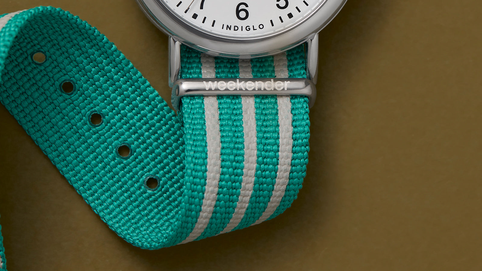 Close-up of the teal-and-white striped fabric strap of the Timex Weekender watch, with “weekender” engraved on the metal loop.