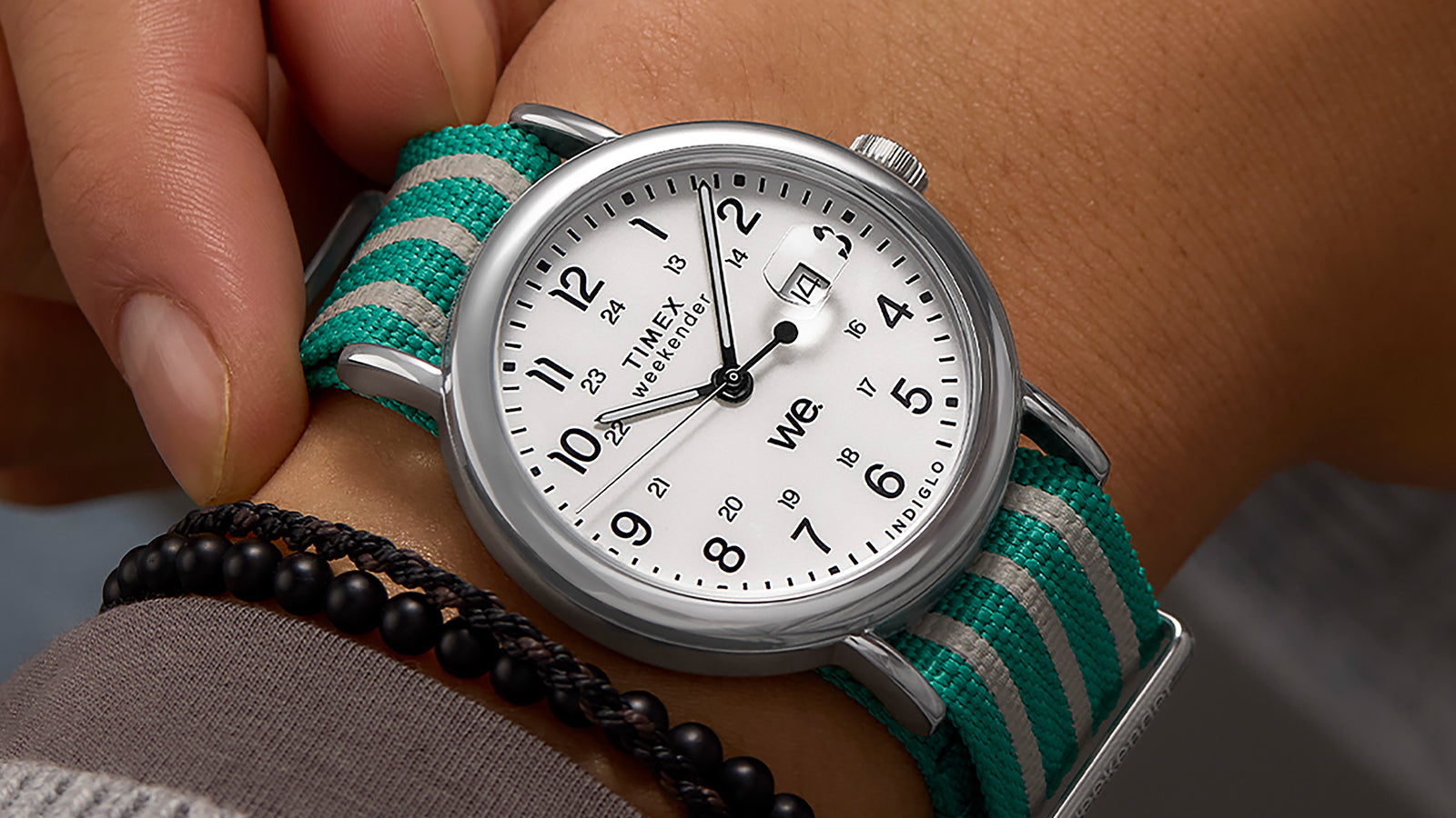 A Timex Weekender watch with teal-and-white striped fabric strap, shown on a wrist alongside a black beaded bracelet.