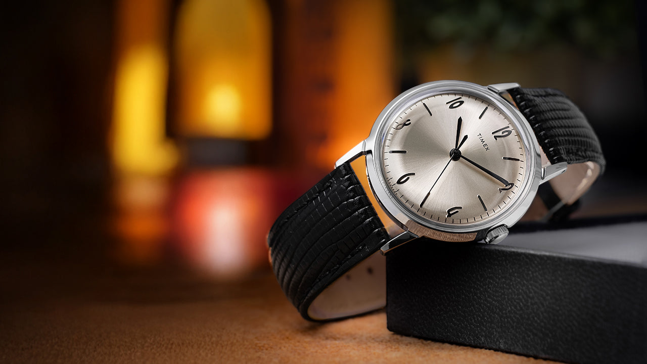 Timex Marlin Hand-Wound watch with silver-tone dial and black leather strap displayed on a box with warm, ambient lighting in the background.