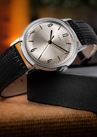 Timex Marlin Hand-Wound watch with silver-tone dial and black leather strap displayed on a box with warm, ambient lighting in the background.
