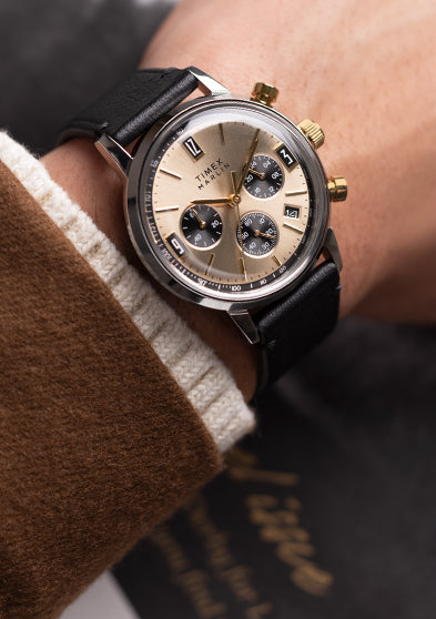 Timex Marlin Chronograph Tachymeter with champagne dial and black leather strap worn on wrist, paired with brown blazer and cream sweater cuff.