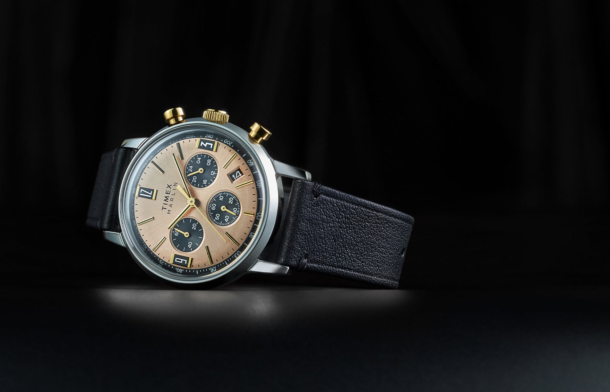 Timex Marlin Chronograph Tachymeter with champagne dial, black subdials, and gold-tone pushers displayed on black leather strap against a dark background.