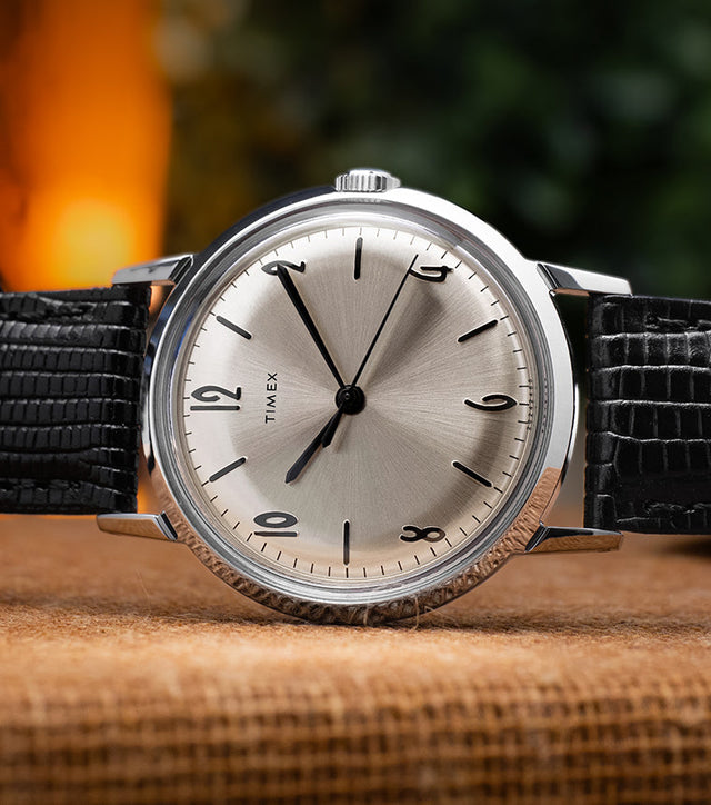Timex Marlin Hand-Wound watch with a silver sunburst dial, black hour markers, and black leather strap resting on a brown fabric surface, warm background lighting.