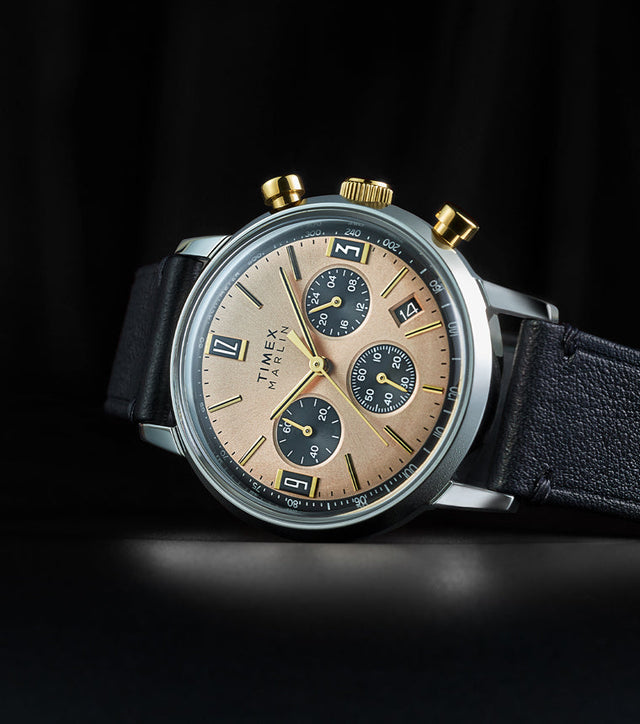 Timex Marlin Chronograph Tachymeter with champagne dial, black subdials, and gold-tone pushers displayed on black leather strap against a dark background.