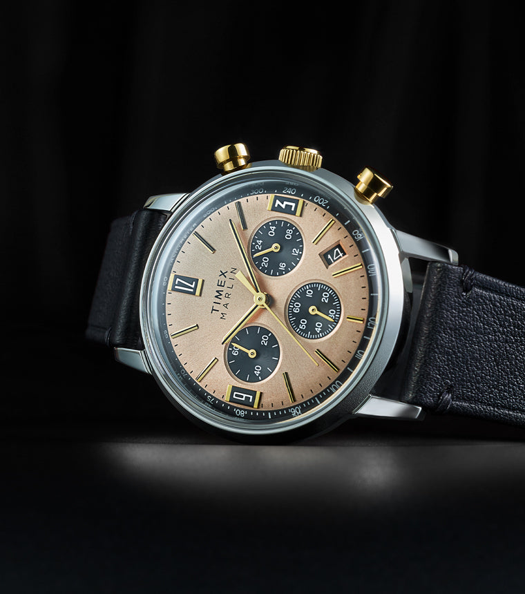 Timex Marlin Chronograph Tachymeter with champagne dial, black subdials, and gold-tone pushers displayed on black leather strap against a dark background.
