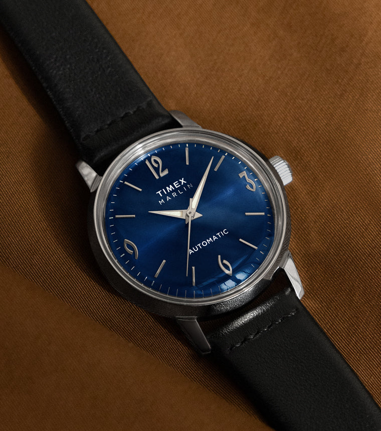 Timex Marlin Automatic watch with deep blue sunray dial and black leather strap resting on a brown fabric surface.