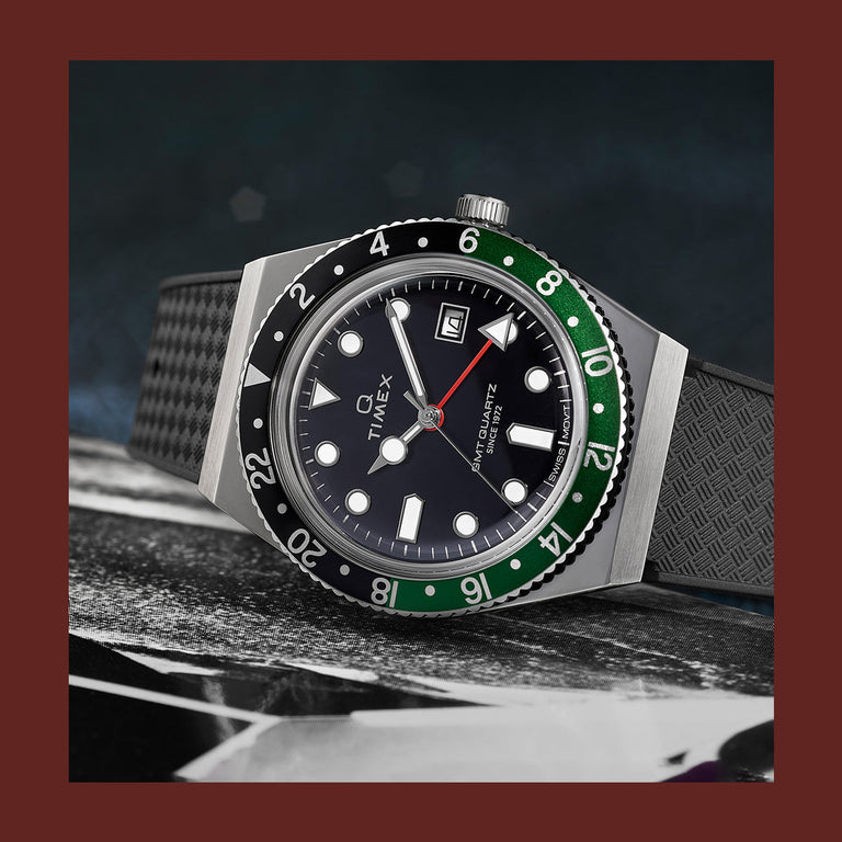 Q Timex GMT watch with a black dial, green and black bezel, red GMT hand, and textured black strap, displayed on a dark surface.