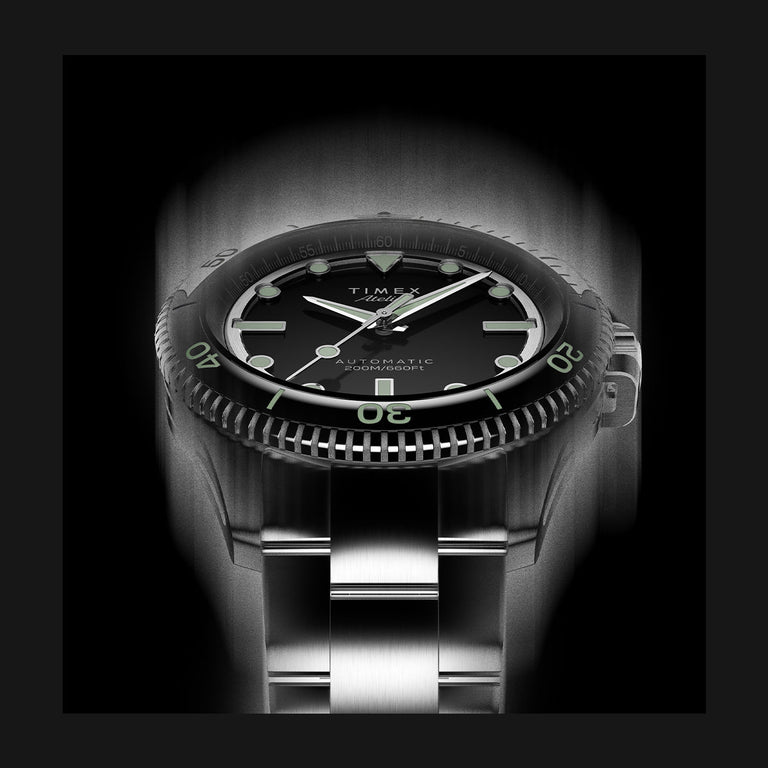 Timex Atelier watch with a black dial, luminous green markers, and stainless steel bracelet, illuminated dramatically in a monochrome lighting effect.