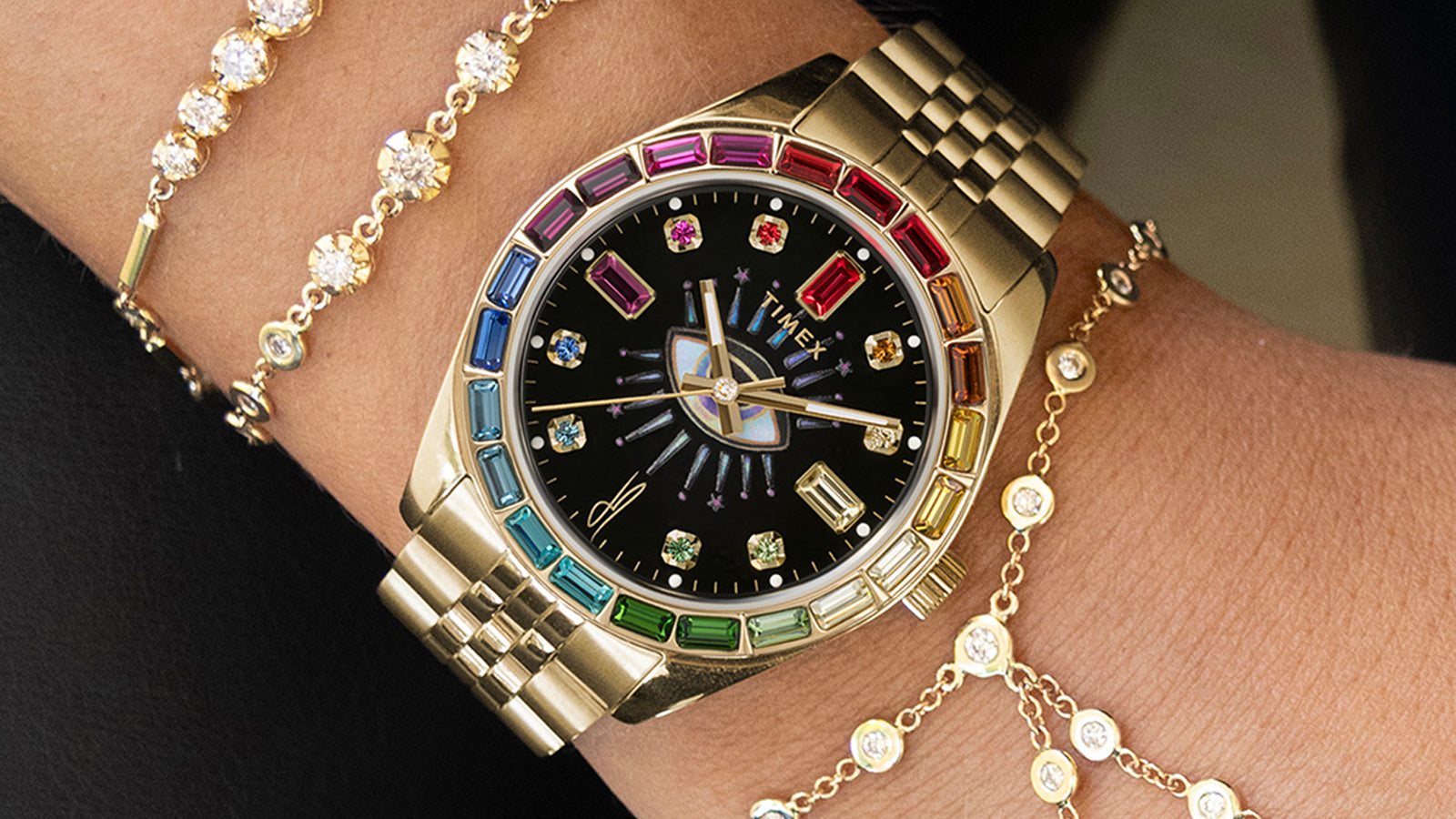 Lifestyle shot of the Timex x Jacquie Aiche “Cosmic Nights” watch worn on the wrist with layered gold and diamond bracelets. The gold case and colorful bezel sparkle under natural light, highlighting its bohemian-luxury style.