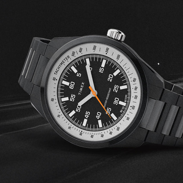 The Huckberry x Timex 1979 Black Max watch angled against a dark, textured background. The black case and bracelet are highlighted, along with the white numerals and orange seconds hand.