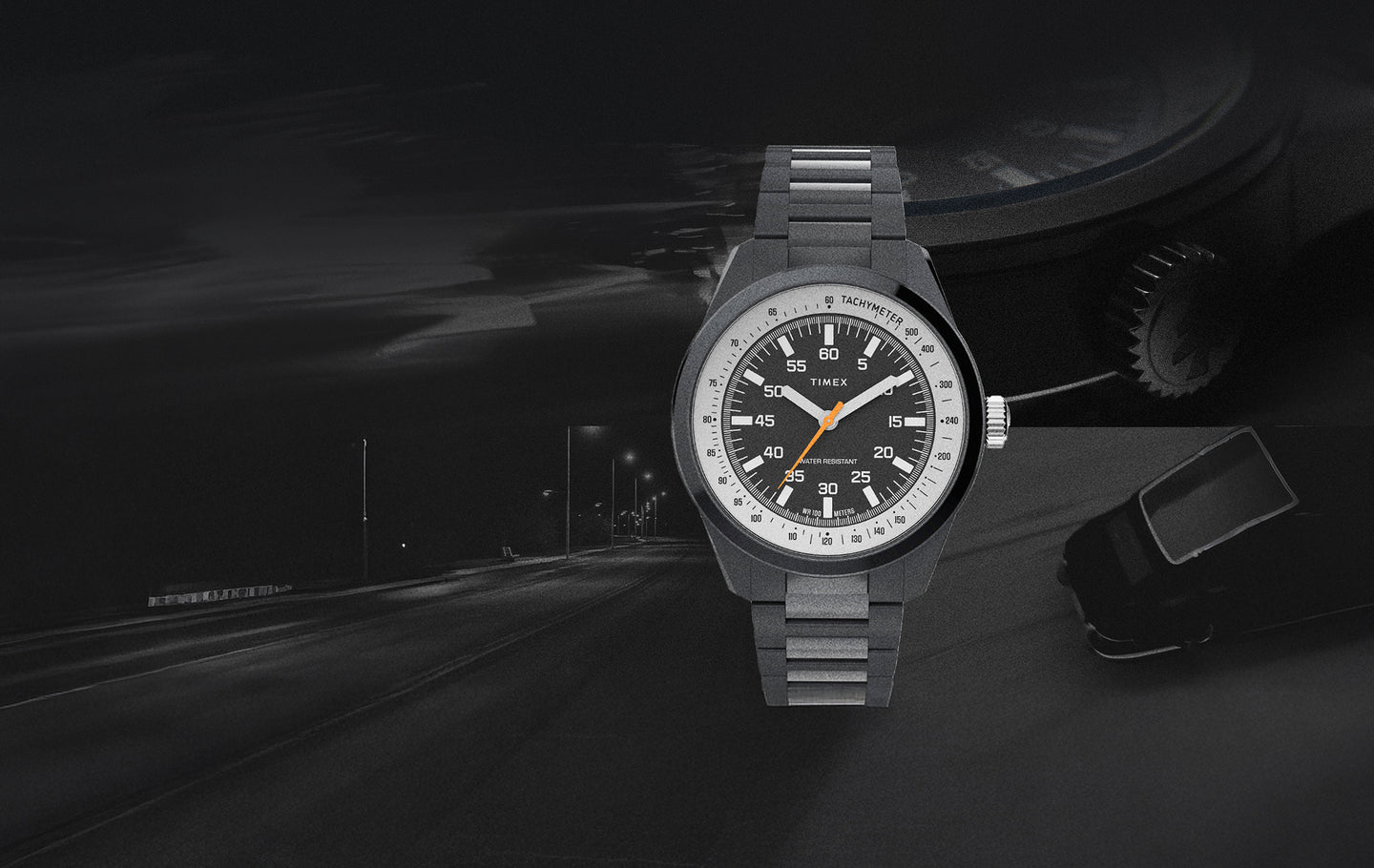 Product image of the Huckberry x Timex 1979 Black Max watch set against a dark, moody background. The watch features a black dial with white markers, an orange seconds hand, and a tachymeter scale, paired with a matte black metal bracelet. A dimly lit nighttime road and a faint outline of a car appear in the background, emphasizing a retro automotive theme.
