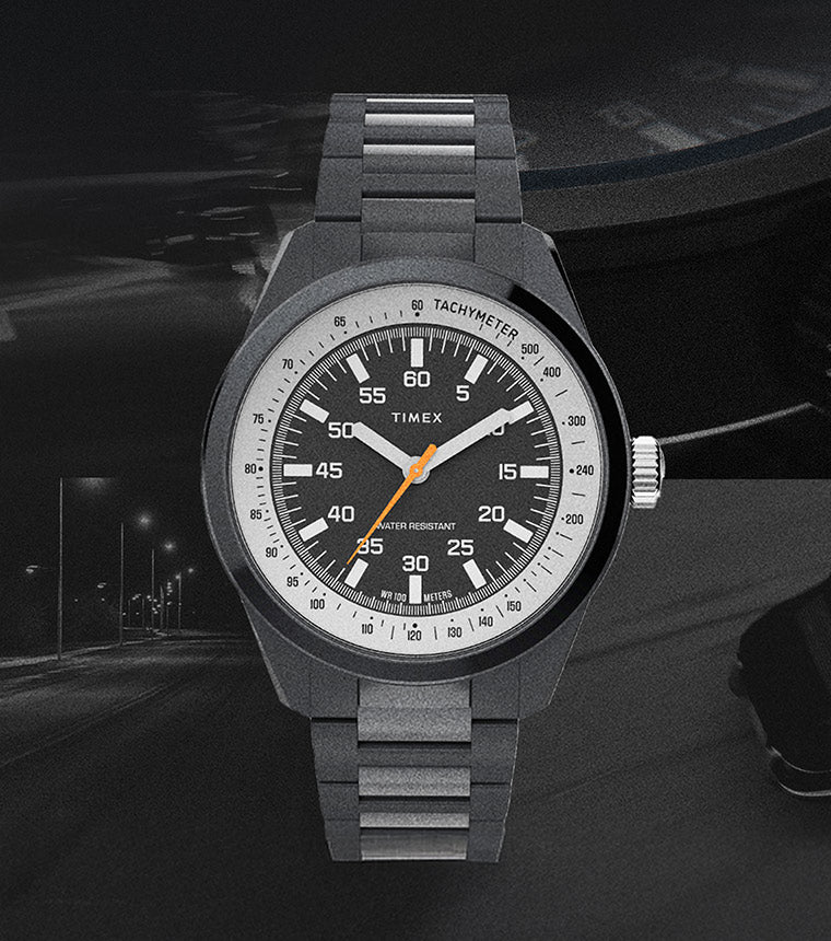 Product image of the Huckberry x Timex 1979 Black Max watch set against a dark, moody background. The watch features a black dial with white markers, an orange seconds hand, and a tachymeter scale, paired with a matte black metal bracelet. A dimly lit nighttime road and a faint outline of a car appear in the background, emphasizing a retro automotive theme.