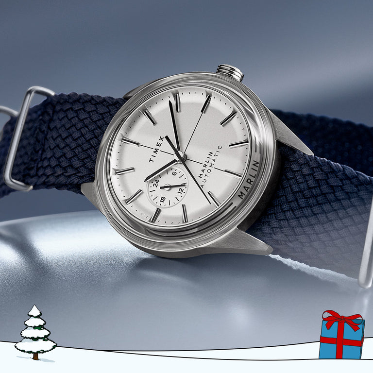 Timex Marlin Jet Automatic watch featuring a brushed stainless steel case, silver dial with black hour markers, and a 24-hour subdial above 6 o’clock. The watch is paired with a textured navy blue fabric strap and displayed against a sleek silver-blue background.