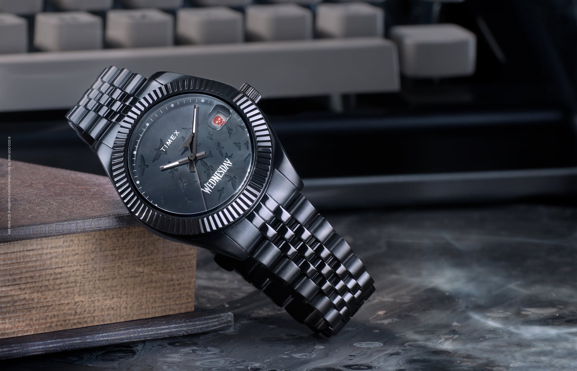 Timex x Wednesday Timex Legacy watch featuring an all-black design with a textured dial, gothic-style “Wednesday” lettering, and a skull icon date window. The watch rests on a vintage book with a dark, moody background, highlighting its mysterious aesthetic.