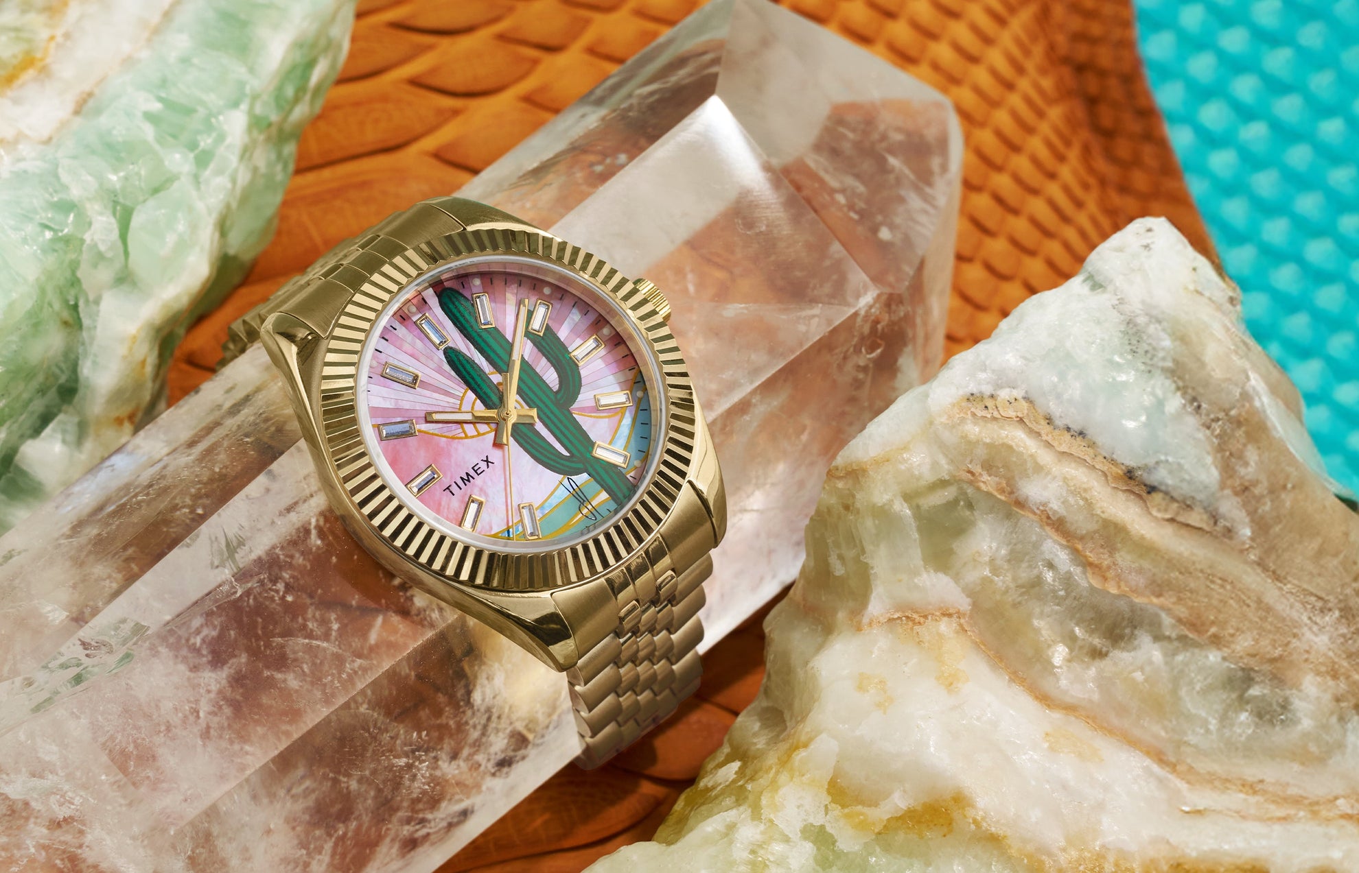 Timex x Jacquie Aiche “Desert Daze” watch with a gold-tone case and bracelet resting on large desert-toned crystals. The dial features a green cactus illustration over a pink mother-of-pearl background with gold hands and hour markers, capturing a Southwestern-inspired aesthetic.