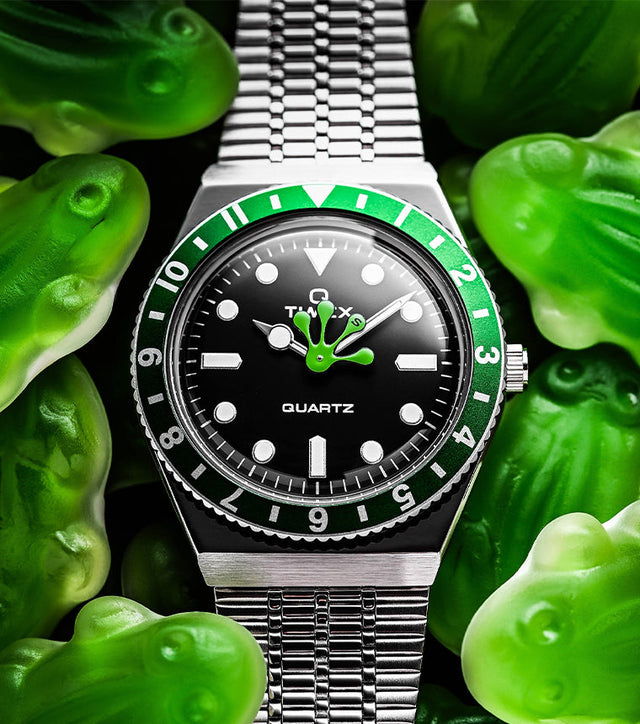 Timex x seconde/seconde/ Q Timex watch with a black dial and green accents surrounded by green gummy frog candies. The hour hand is playfully shaped like a frog’s foot, blending humor and design creativity in seconde/seconde/’s signature style.