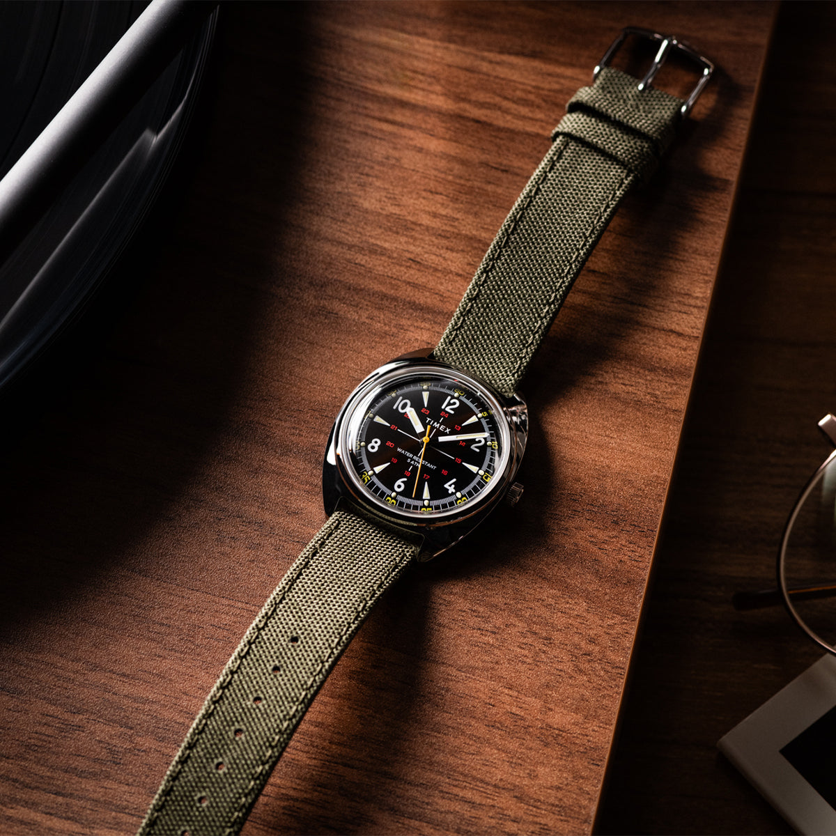 Overhead shot of the Timex x Bespoke Post watch on a wooden surface, showcasing its polished silver-tone case, black dial with red and yellow details, and green woven strap. Warm lighting emphasizes its vintage-inspired design.