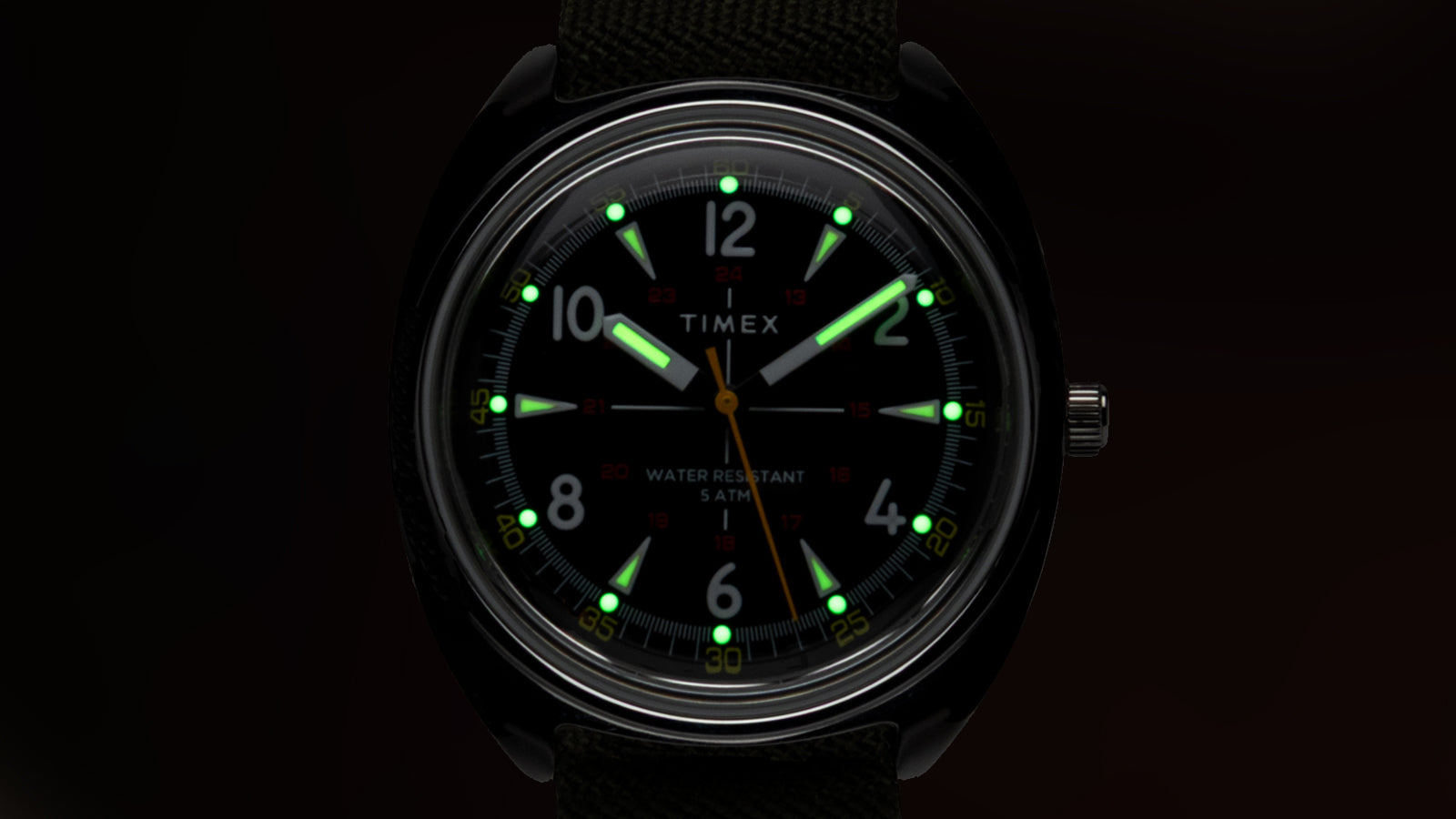 Front view of the Timex x Bespoke Post watch with luminescent hands and markers glowing in green. The black dial and bright lume contrast create a clear and functional nighttime display.