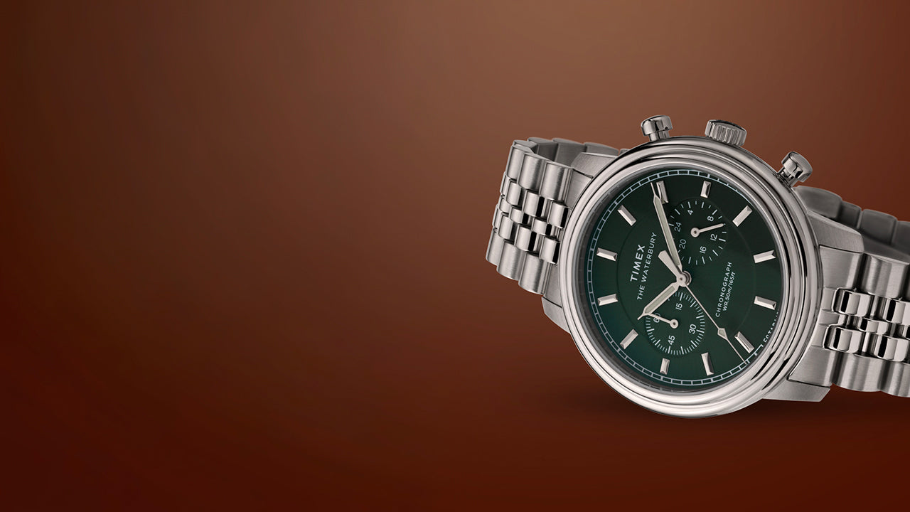 Timex Waterbury Metropolitan Chronograph watch featuring deep green dial, chronograph sub-dials, and polished stainless steel bracelet on a brown backdrop.
