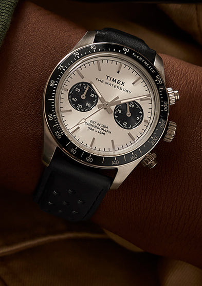 Timex Waterbury Heritage Chronograph on wrist, showing silver-tone dial, black sub-dials, and black perforated leather strap paired with a green sweater.